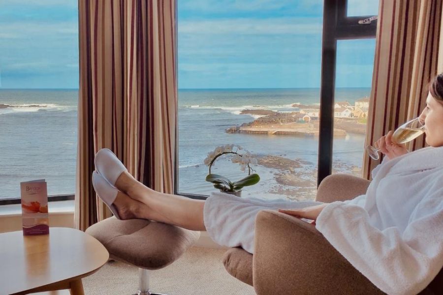 Een vrouw die champagne aan het drinken is in haar witte badjas en staart naar buiten waar zij uitzicht heeft op de zee